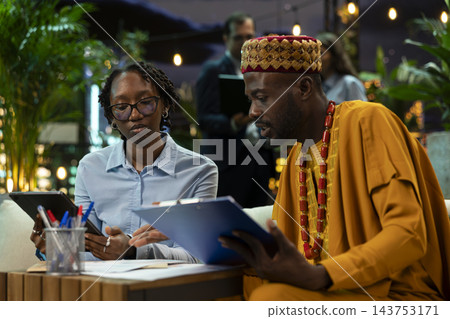 Business partners compare information on tablet and printed documents during discussion at night. Professionals crosschecking data between device and paperwork on clipboard in rooftop workspace Business partners compare information on tablet and printed documents during discussion at night. Professionals crosschecking data between device and paperwork on clipboard in rooftop workspace 143753171