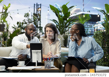 Businessman points to tablet screen during rooftop lounge workspace presentation with coworkers. Company team analyzing project information on device during meeting on rooftop terrace Businessman points to tablet screen during rooftop lounge workspace presentation with coworkers. Company team analyzing project information on device during meeting on rooftop terrace 143753172