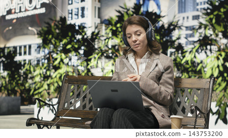 Businesswoman works remotely from park bench, having video conference on laptop. Woman wearing headphones communicates with colleagues online, enjoying flexible outdoor workplace Businesswoman works remotely from park bench, having video conference on laptop. Woman wearing headphones communicates with colleagues online, enjoying flexible outdoor workplace 143753200