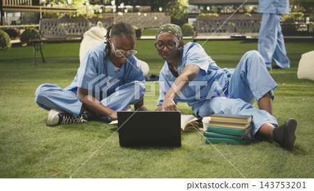 Women students team in blue scrubs work together with laptop and textbooks, group study on campus grass with stacked books. Outdoor learning and med school study session. Women students team in blue scrubs work together with laptop and textbooks, group study on campus grass with stacked books. Outdoor learning and med school study session. 143753201