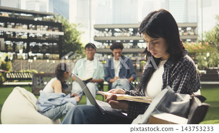 Focused woman student working on laptop while sitting on bench in modern campus with stacks of books and coffee cup nearby. Independent study session outdoors. 143753233