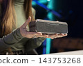 Wireless speaker, woman and product presentation as she holds audio device close to camera. Technology review, demonstration and electronics marketing support an informative product showcase. 143753268
