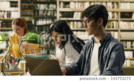 Laptop, student and studying as young man with glasses works on a computer beside fellow readers. Research, digital learning and academic productivity support coursework in a library. Laptop, student and studying as young man with glasses works on a computer beside fellow readers. Research, digital learning and academic productivity support coursework in a library. 143753273