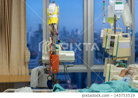Infusion bags hang beside electronic medical pumps near hospital bed green bedding patient partly visible while large window reveals distant city view cool evening light 143753519