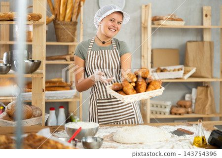Smiling female baker in uniform stands behind counter in small private bakery offering croissants Smiling female baker in uniform stands behind counter in small private bakery offering croissants 143754396