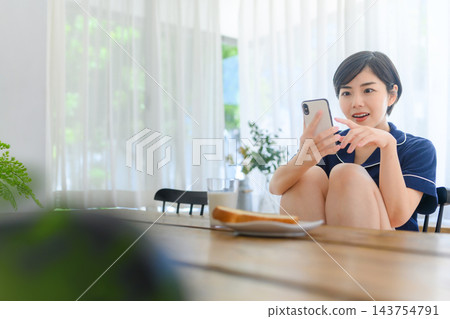 一位年輕女子在餐桌上用智慧型手機吃早餐。 一位年輕女子在餐桌上用智慧型手機吃早餐。 143754791