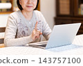 A middle-aged or senior woman holding up her index finger while using a computer in her living room at home. 143757100