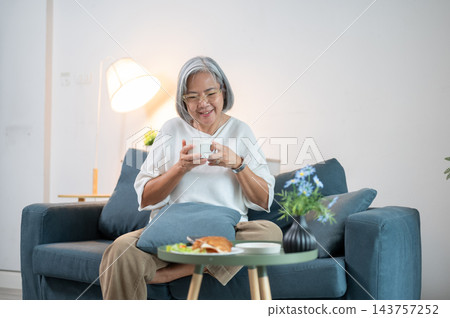 Senior Asian woman smiling while holding coffee cup on living room sofa. Senior Asian woman smiling while holding coffee cup on living room sofa. 143757252