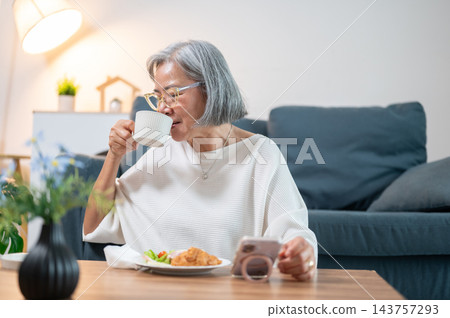 Senior Asian Woman Drinking Coffee and Looking at Smartphone at Home Senior Asian Woman Drinking Coffee and Looking at Smartphone at Home 143757293