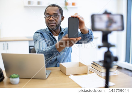 A man presents a smartphone at a desk while recording a video, with a laptop, open parcel box, stacked books, and camera equipment in a bright workspace A man presents a smartphone at a desk while recording a video, with a laptop, open parcel box, stacked books, and camera equipment in a bright workspace 143759044