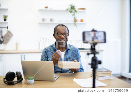A smiling man speaks to a smartphone camera at a kitchen table, with a laptop, headphones, stacked books, and a small box in a bright home kitchen A smiling man speaks to a smartphone camera at a kitchen table, with a laptop, headphones, stacked books, and a small box in a bright home kitchen 143759059