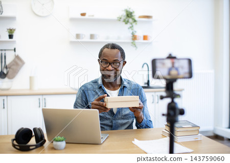 An African American man presents a small cardboard package to a smartphone camera from a bright home workspace, with a laptop and books nearby An African American man presents a small cardboard package to a smartphone camera from a bright home workspace, with a laptop and books nearby 143759060