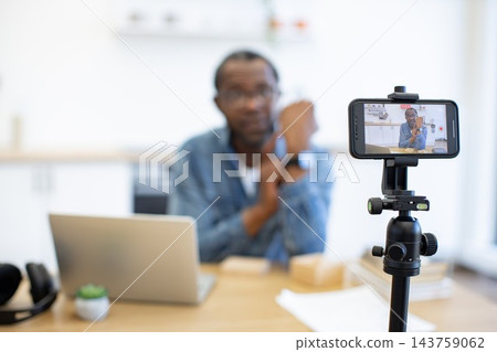 A man records himself with a smartphone on a tripod at a desk, alongside a laptop, headphones, papers, and a small plant in a bright workspace A man records himself with a smartphone on a tripod at a desk, alongside a laptop, headphones, papers, and a small plant in a bright workspace 143759062