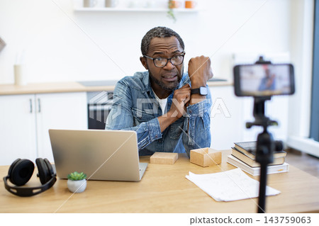 A man presents a smartwatch to a phone camera at a home desk, with a laptop, headphones, books, and shipping boxes arranged around his workspace A man presents a smartwatch to a phone camera at a home desk, with a laptop, headphones, books, and shipping boxes arranged around his workspace 143759063