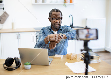 A man checks his smartwatch while recording a presentation at a home kitchen table, with a laptop, headphones, books and papers nearby 143759064