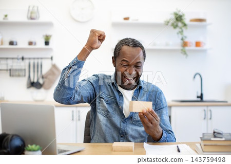 A joyful African American man raises a fist while holding a small parcel at his desk, with a laptop, paperwork, and a bright kitchen behind him 143759498