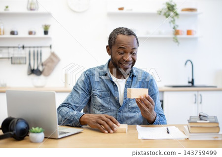 An African American man smiles while examining a small cardboard package at his desk, with a laptop, headphones, notebook, and books in a bright workspace 143759499