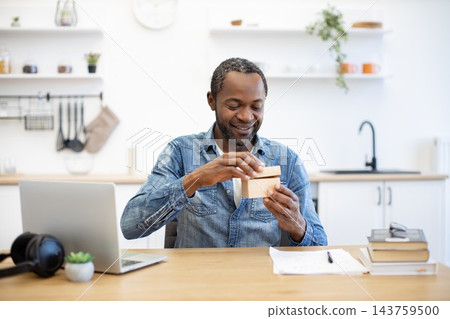 A smiling man opens a small cardboard box at his desk, with a laptop, headphones, books, and a bright modern kitchen in the background A smiling man opens a small cardboard box at his desk, with a laptop, headphones, books, and a bright modern kitchen in the background 143759500