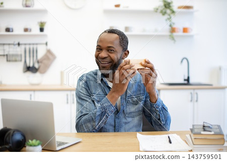 A cheerful man pauses with a sandwich at a wooden table, laptop and notes nearby, taking a relaxed work break in a bright home kitchen 143759501