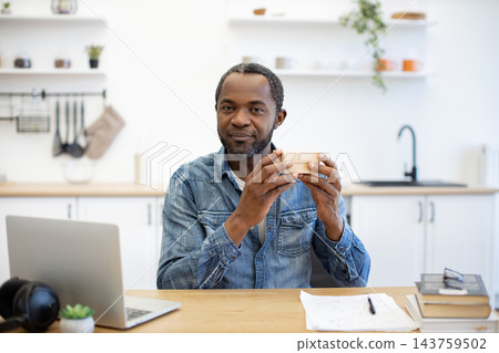 A bearded man sits at a kitchen table holding a small wooden object, with a laptop, handwritten notes, books, and glasses nearby in a bright room 143759502