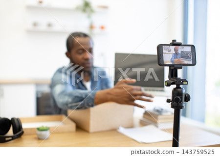 A creator demonstrates a cardboard package at a desk, filming with a smartphone on a tripod beside a laptop in a bright home workspace 143759525