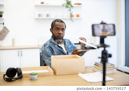 A content creator records a laptop unboxing at a bright home workspace, with cardboard packaging, books, headphones, and a phone mounted on a tripod 143759527