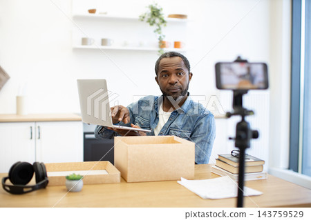 An African American man works at a desk with a laptop and shipping boxes while a smartphone on a tripod records his home workspace for online content 143759529