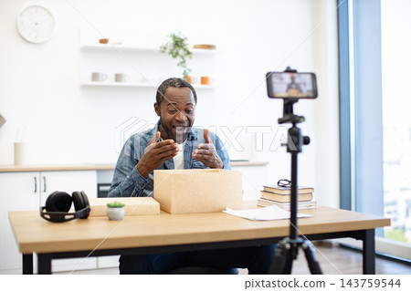 An African American man gestures toward an open cardboard package while recording a video at a bright home desk with a smartphone on a tripod An African American man gestures toward an open cardboard package while recording a video at a bright home desk with a smartphone on a tripod 143759544