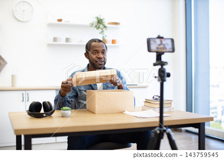 An African American man opens a cardboard package at his desk while recording a presentation with a smartphone in a bright home workspace 143759545