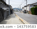 日本住宅區白天的寧靜街景，電線桿和電線沿著道路和人行道延伸，兩旁是獨立式住宅和老舊建築，遠處可以看到山脈。 143759583