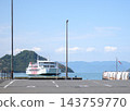 一艘大型白色渡輪漂浮在平靜的海面上，背景是藍天和綠色的島嶼，旁邊是日本港口的碼頭和瀝青停車場。 143759770