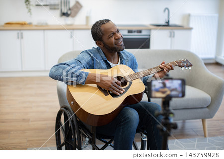 An African American musician plays acoustic guitar from his wheelchair in a bright home, while a smartphone on a tripod records the performance An African American musician plays acoustic guitar from his wheelchair in a bright home, while a smartphone on a tripod records the performance 143759928