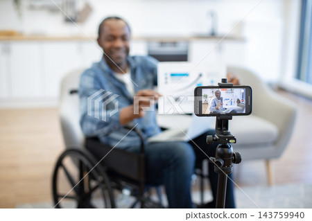 A man in a wheelchair demonstrates a boxed item while filming himself with a smartphone mounted on a tripod in a bright, modern indoor setting 143759940