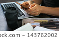 Hands using smartphone beside laptop on desk with blurred coffee cup, printed photos, and pen, suggesting online browsing, message checking, digital work, and casual planning in modern workspace 143762002
