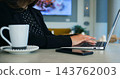 Businesswoman working on laptop computer while sitting near warm beverage cup in cafe setting. Female employee types attentively on modern device during productive remote office workday. 143762003