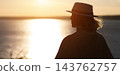 Woman stands quietly beside glowing water, wearing fedora and looking toward sunset. Dark silhouette, relaxed posture, and warm reflected sunlight create serene contemplative mood. 143762757