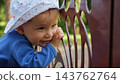 Joyful Infant Smiles Near Metal Railing In Tranquil Summer Garden Setting. Curious Little Child In Patterned Sun Hat Gazes Away, Showing Pure Happiness Amid Blurred Trees And Sunlight Outdoors. 143762764