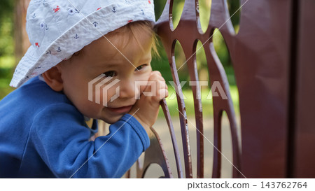 Joyful Infant Smiles Near Metal Railing In Tranquil Summer Garden Setting. Curious Little Child In Patterned Sun Hat Gazes Away, Showing Pure Happiness Amid Blurred Trees And Sunlight Outdoors. Joyful Infant Smiles Near Metal Railing In Tranquil Summer Garden Setting. Curious Little Child In Patterned Sun Hat Gazes Away, Showing Pure Happiness Amid Blurred Trees And Sunlight Outdoors. 143762764