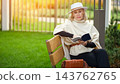 Older woman reads book on bench in bright garden. Elegant reader holds open pages with focused calm, presenting retirement leisure through relaxed outdoor reading and polished personal style. 143762765