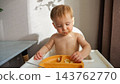 Shirtless Toddler Explores Fruit Slices Inside Suction Plate While Seated In White Highchair. Curious Little Child Looks Down At Healthy Food Snack, Enjoying Calm Morning Meal With Dirty Face. 143762770