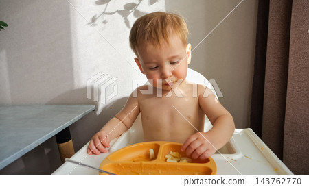 Shirtless Toddler Explores Fruit Slices Inside Suction Plate While Seated In White Highchair. Curious Little Child Looks Down At Healthy Food Snack, Enjoying Calm Morning Meal With Dirty Face. Shirtless Toddler Explores Fruit Slices Inside Suction Plate While Seated In White Highchair. Curious Little Child Looks Down At Healthy Food Snack, Enjoying Calm Morning Meal With Dirty Face. 143762770