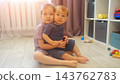 Smiling Girl Holds Little Brother Closely Near Sunlit Window In Cozy Playroom. Tender Sister Supports Barefoot Toddler Sibling With Gentle Care, Expressing Innocent Love During Play Session. 143762783