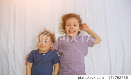 Two Cheerful Siblings Rest Together On White Bed, Smiling Joyfully During Bedtime Routine; Happy Girl Embraces Younger Toddler Brother While Staying Awake Under Bright Bedroom Illumination. 143762786