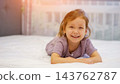 Happy Young Girl Rests Peacefully On Comfortable Bed Inside Cozy Modern Bedroom. Delighted Child Smiles Toward Camera While Relaxing On Clean White Sheets During Pleasant Sunny Morning At Home. 143762787