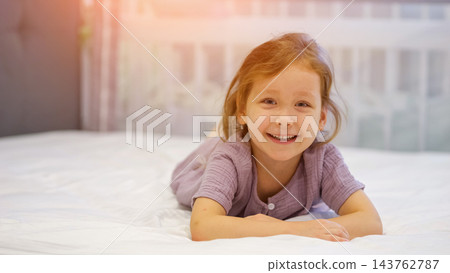 Happy Young Girl Rests Peacefully On Comfortable Bed Inside Cozy Modern Bedroom. Delighted Child Smiles Toward Camera While Relaxing On Clean White Sheets During Pleasant Sunny Morning At Home. 143762787