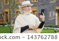 Woman poses with smartphone in outdoor portrait near modern building. She wears white hat and cream cape, and her smiling expression gives personal profile image stylish, elegant feel. 143762788