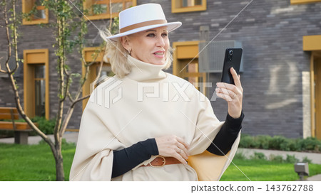 Woman poses with smartphone in outdoor portrait near modern building. She wears white hat and cream cape, and her smiling expression gives personal profile image stylish, elegant feel. Woman poses with smartphone in outdoor portrait near modern building. She wears white hat and cream cape, and her smiling expression gives personal profile image stylish, elegant feel. 143762788
