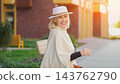 Smiling woman poses outdoors in white hat and cape. Woman stands beside sunlit walkway, facing camera with relaxed confidence in stylish portrait near building and soft garden backdrop. 143762790