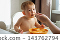 Shirtless Toddler Eats Puree Spoon From Caregiver Hand While Sitting In White High Chair Indoors. Young Child Tastes Baby Food During Picky Eating Mealtime Routine Beside Yellow Plate. 143762794