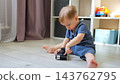 Toddler Sits On Floor Examining Vintage Camera With Curious Intent. Barefoot Infant Plays Carefully Indoors Beside Toy Shelf, Exploring Retro Mechanical Device With Quiet, Focused Expression. 143762795